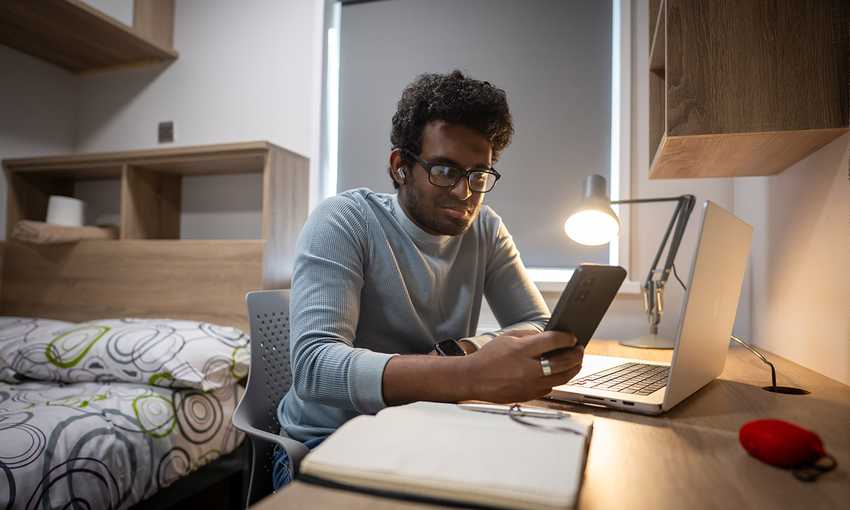 A man seated at a desk, working on a laptop while holding a cell phone in his hand.