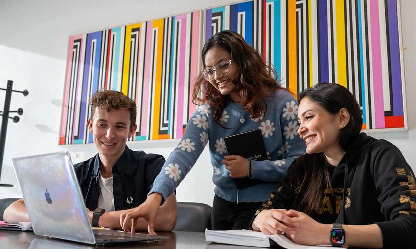 Three students collaborating on a laptop in a modern office setting, focused on their project.