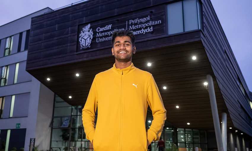 A man wearing a yellow jacket stands confidently in front of a Cardiff Met building, showcasing a vibrant urban scene.