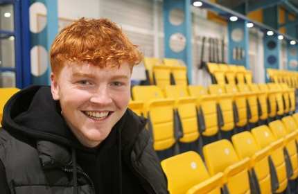 A young man with red hair smiles brightly, standing in front of vibrant yellow seats.