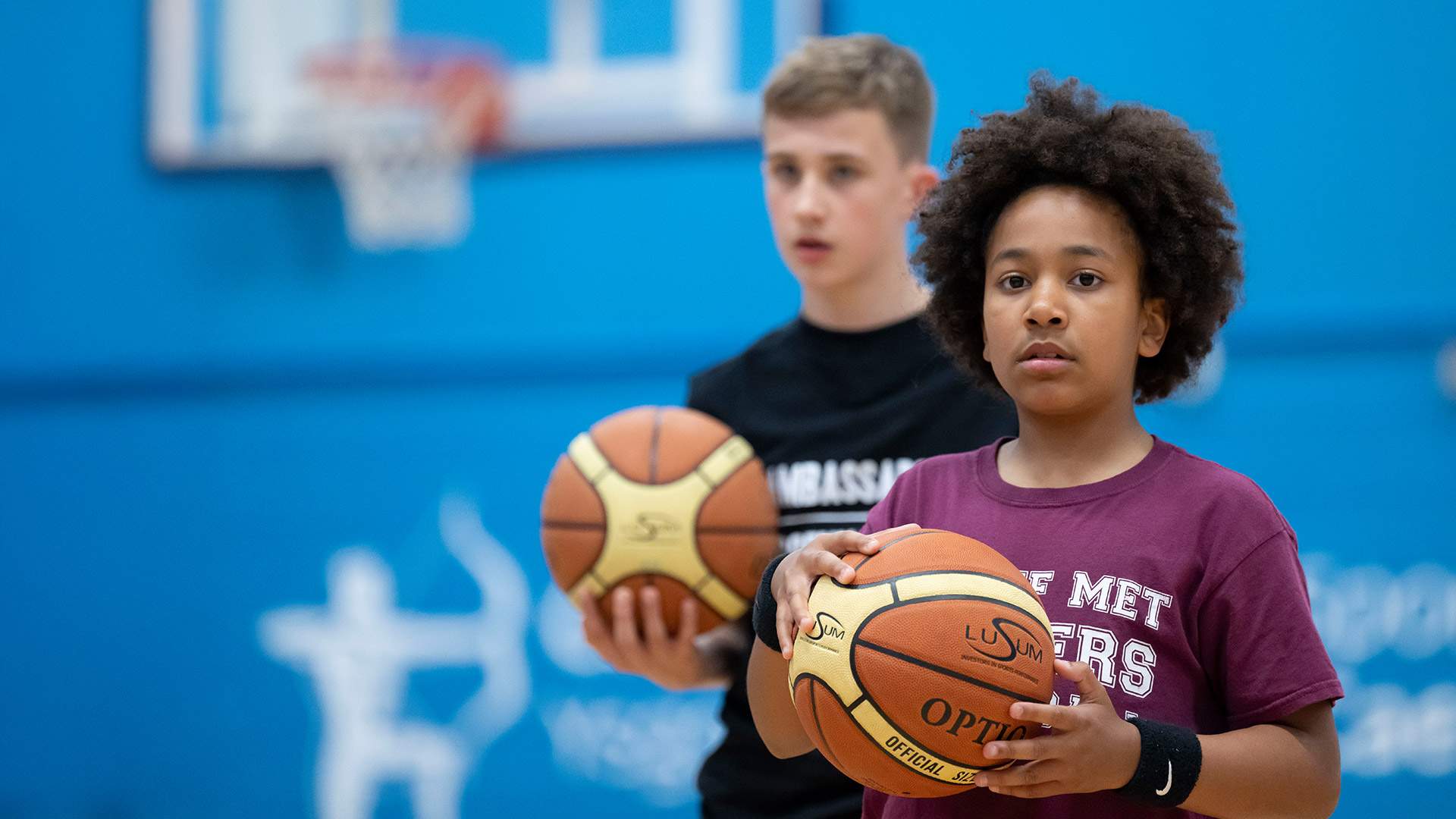 Two young boys stand in a gym, each holding a basketball, ready to play and enjoy their time together.