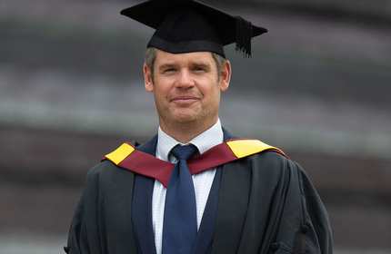 A man wearing a graduation gown and tie, smiling proudly at his graduation ceremony.
