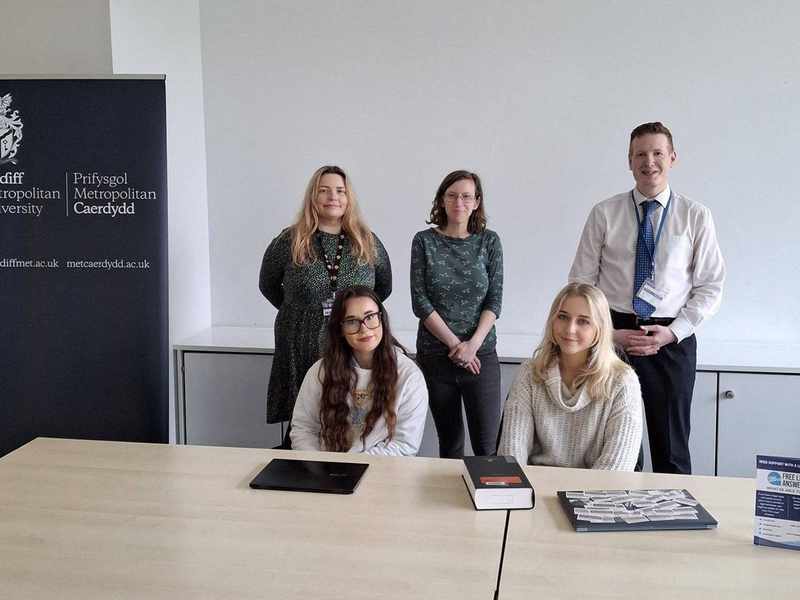 Five people gathered around a table. On the table are two laptop computers and a hardback book.