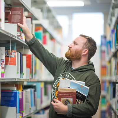 A man with a beard in a green hoodie stands in a library aisle, holding several books in one arm and reaching up to grab another book from a shelf. The library is well-lit with various colorful books on the shelves.