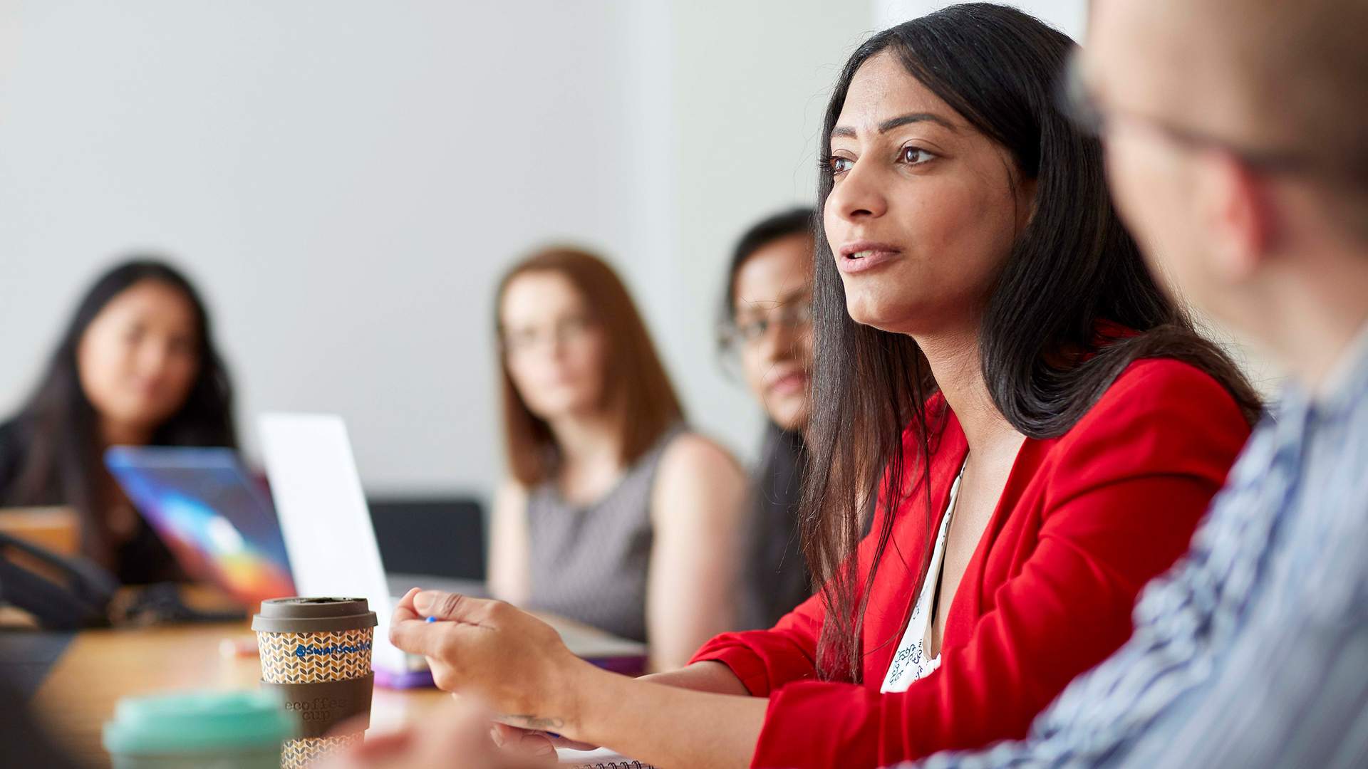 A woman in a red blazer speaks during a meeting, surrounded by colleagues sitting at a conference table. Some have laptops open, and a coffee cup is visible in the foreground. The setting is a well-lit office room.