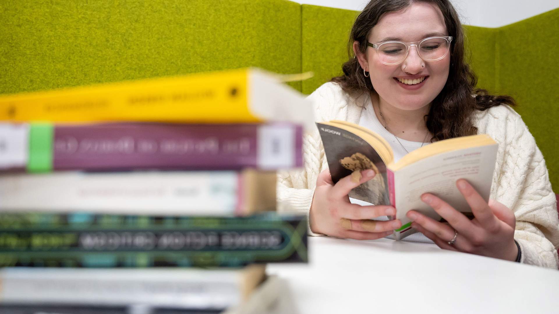 A person with glasses is smiling while reading a book. They are seated at a table with a stack of books in the foreground. The background features green upholstered panels.
