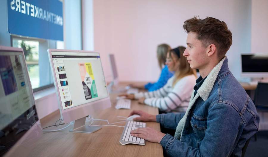 A person sat at a desk in front of a Mac computer. In the background are two other people also using computers.