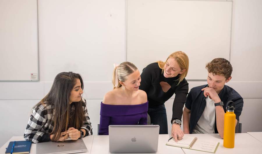 Three people sit alongside each other at a table with laptop computers and notebooks. Behind them another person leans in, pointing to an open notebook.