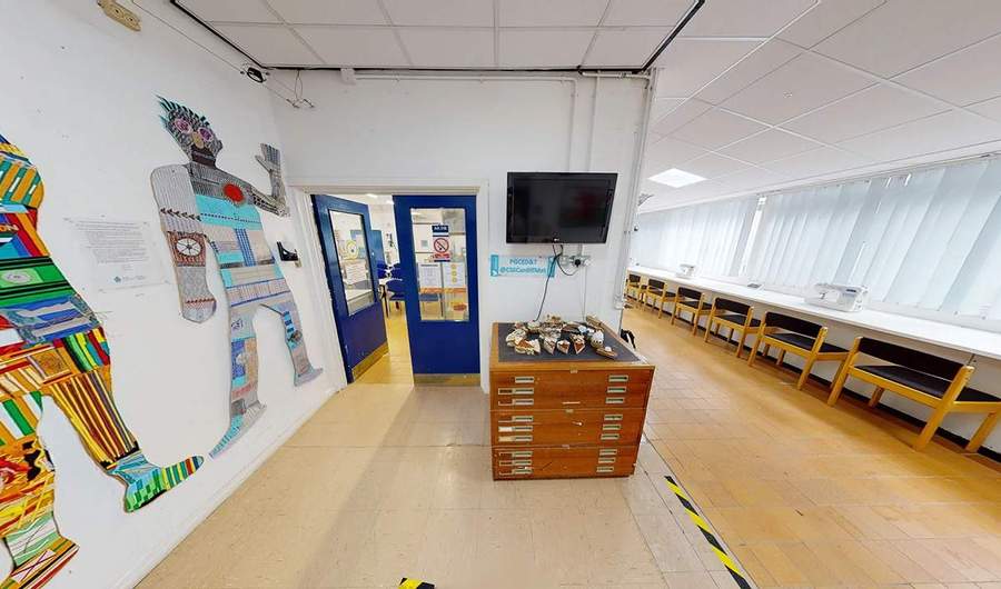 A corridor outside a room with desks and chairs. Mounted on the wall are stylised figures made from different materials.