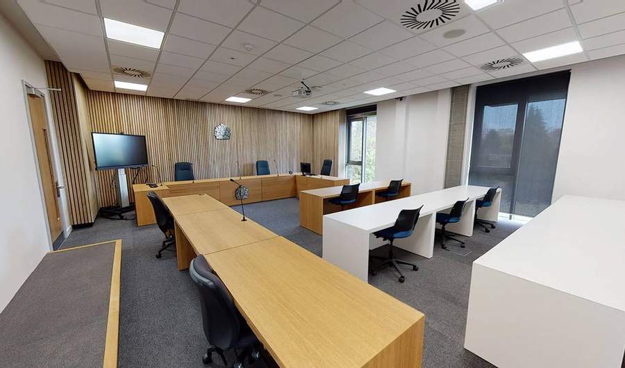 A large room with desks and chairs arranged in rows. At the far end is a long desk with a crown symbol mounted on it.