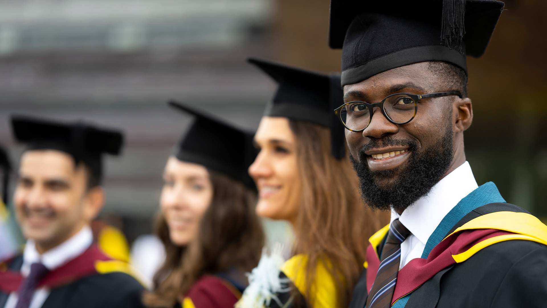 A graduate in cap and gown joyfully holds up a Cardiff Metropolitan University sign. The background is blurred, highlighting his smile and the sign.