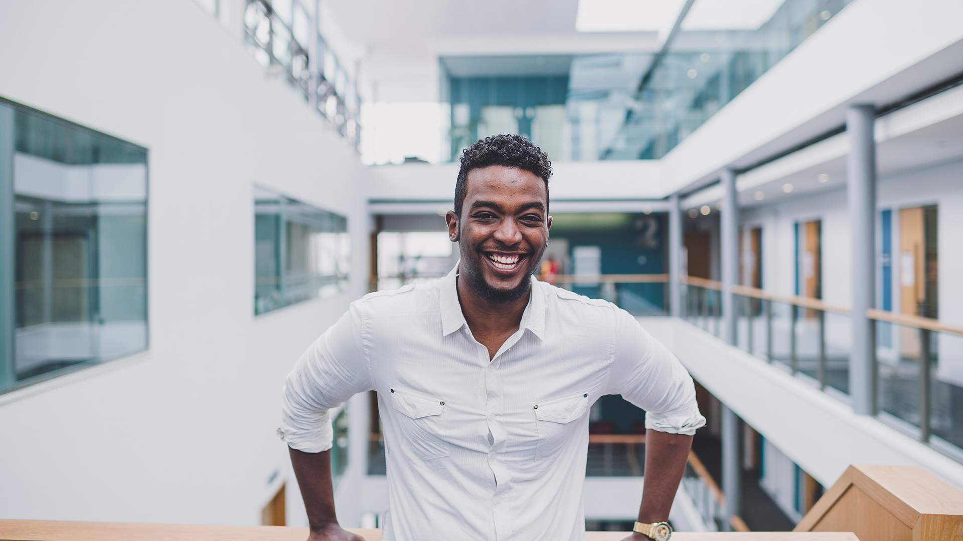 A person in a yellow jacket stands smiling in front of Cardiff Metropolitan University, with the buildings sign visible in the background against a cloudy sky.