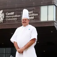 A man in a chef's uniform stands outside a building, smiling and holding a cooking utensil.