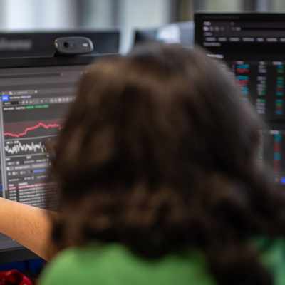Two men are standing in a modern building with large windows. One is leaning against a wooden railing, wearing a red shirt and cap, while the other stands facing him, wearing a white shirt and watch. They are smiling and engaged in conversation.