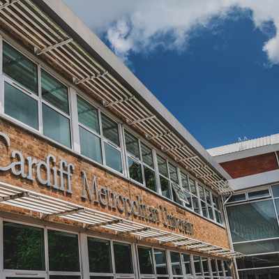 A building with large windows featuring the sign Cardiff Metropolitan University on a brick wall. A blue sky with clouds is visible above.