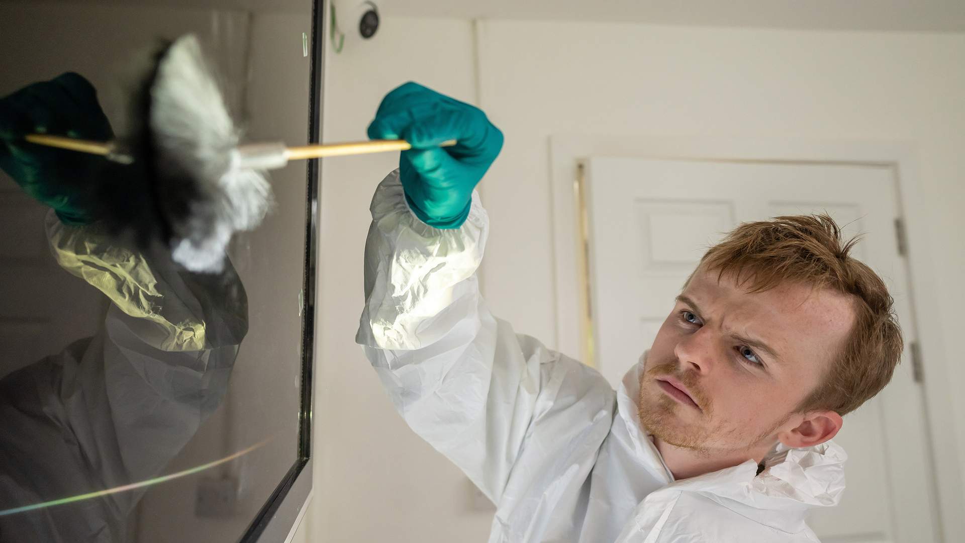 A person wearing a protective suit and teal gloves analyzes fingerprints on a reflective surface, marked with green evidence tags. The image is brightly lit, casting rainbow-like reflections.