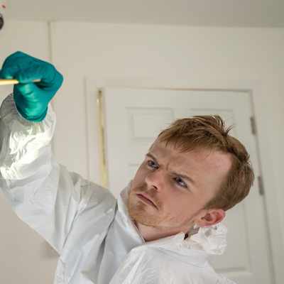 A person wearing a protective suit and teal gloves analyzes fingerprints on a reflective surface, marked with green evidence tags. The image is brightly lit, casting rainbow-like reflections.
