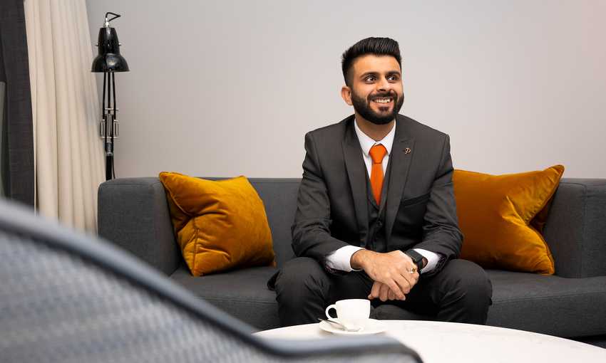 A man wearing a dark suit with an orange tie stands smiling in a modern office lobby with wooden floors and open double glass doors in the background. An orchid is visible on a table to the left.