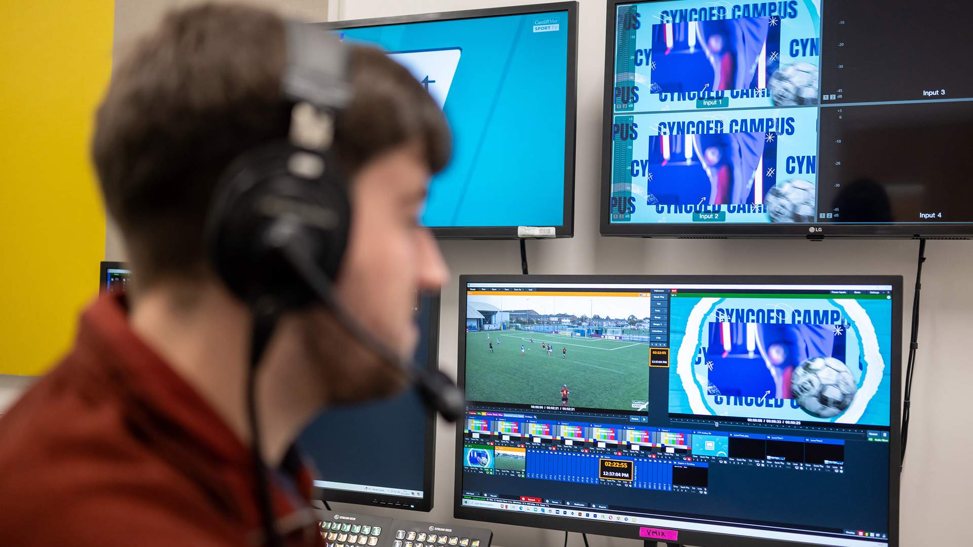 A person in headphones watches a football match on several monitors.