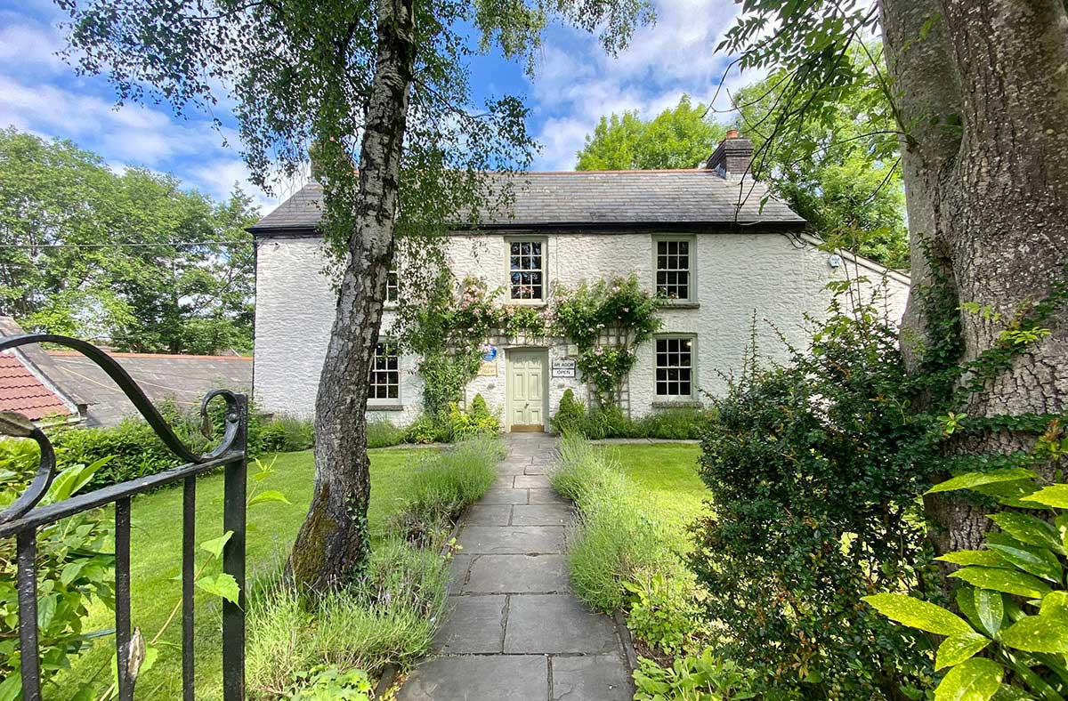 A stone path leads to a white two-story cottage with a slate roof, central door, and green garden, surrounded by trees and plants under a partly cloudy sky.