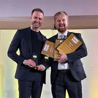 Two men in suits proudly holding awards on stage, celebrating their achievements in a formal event setting.