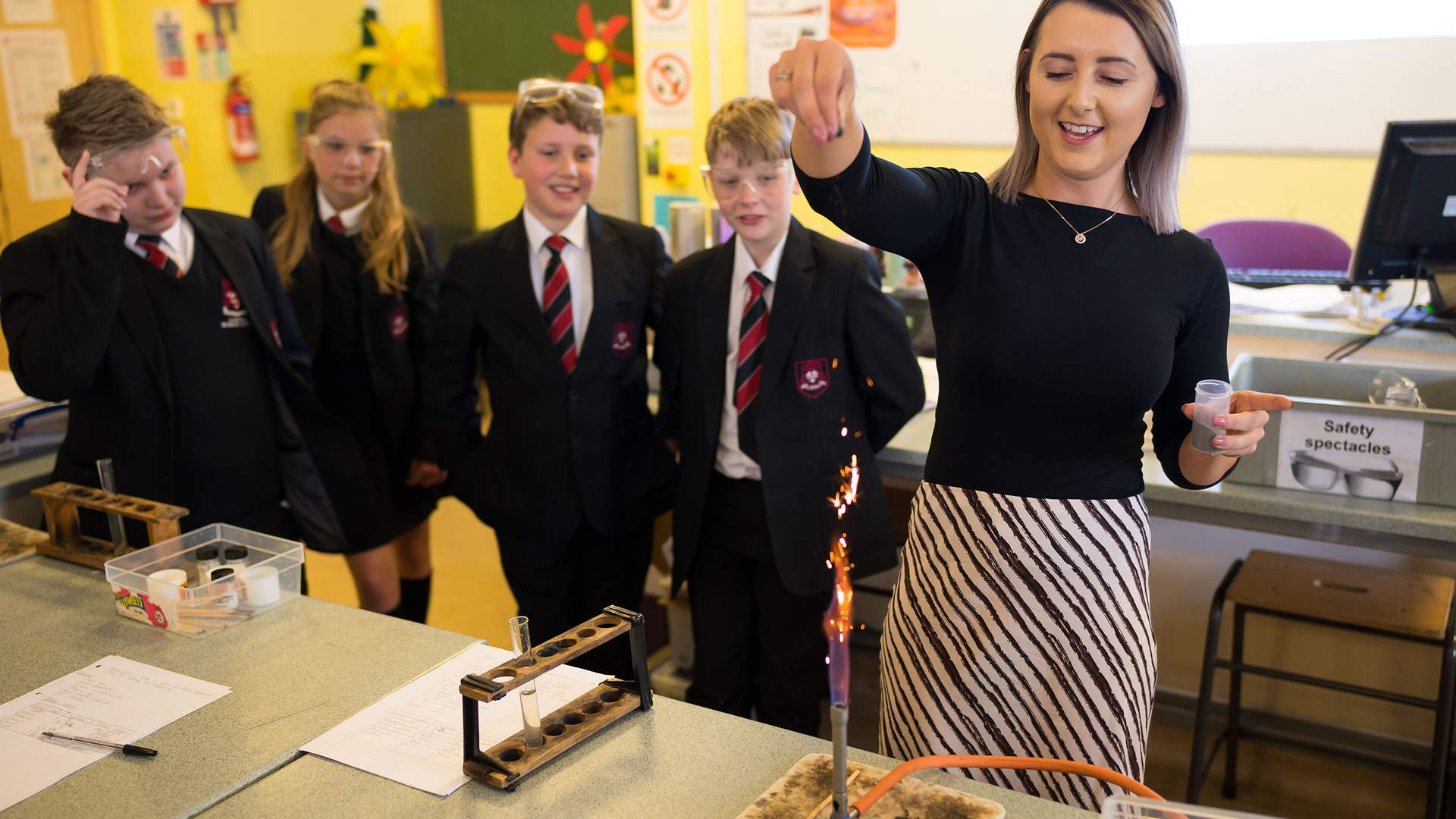 A teacher conducts a chemistry experiment with a Bunsen burner, producing sparks, while four students in school uniforms watch attentively in a classroom. Safety equipment is visible on the table.