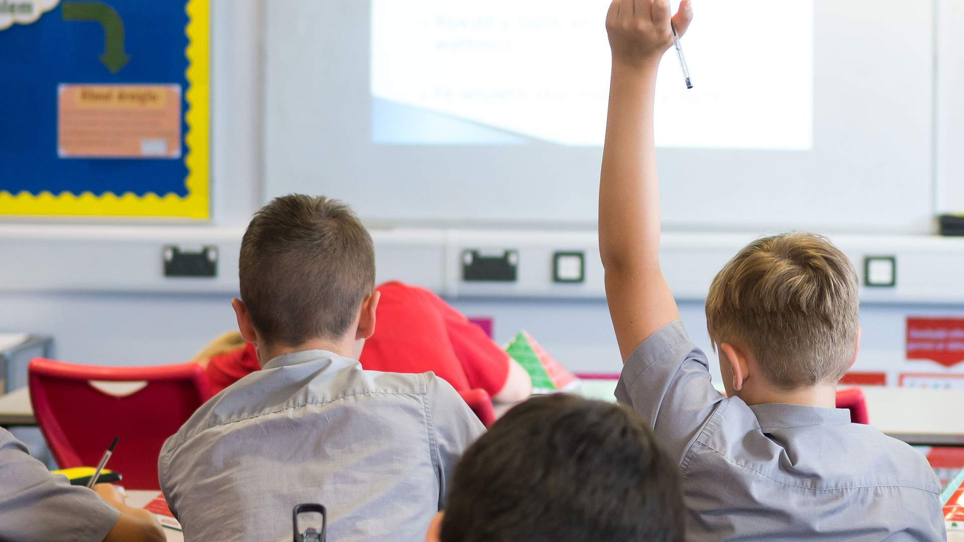 Two pupils are sitting in a classroom. One pupil has his hand raised.