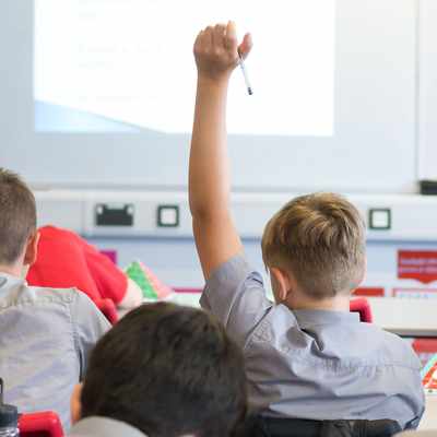 Two pupils are sitting in a classroom. One pupil has his hand raised.