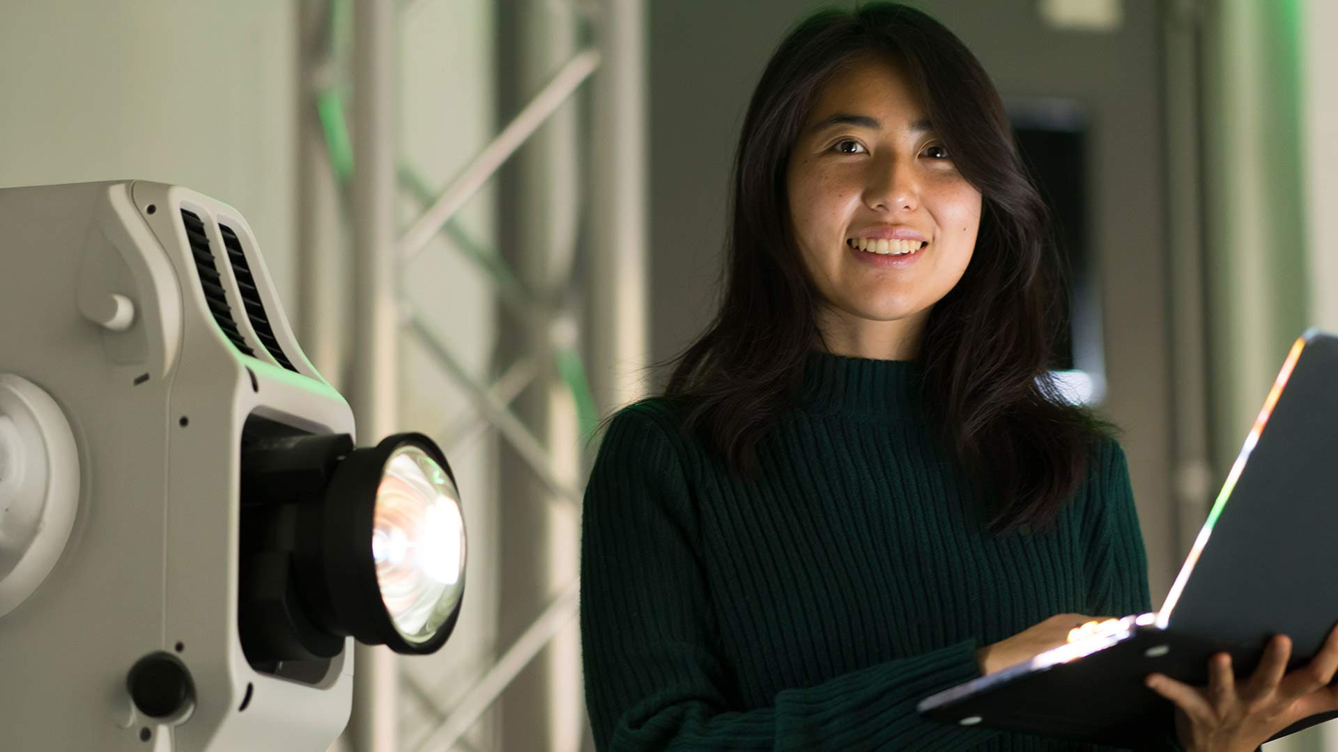 A woman with a laptop in hand is positioned in front of a gentle light, highlighting her concentration and intent.