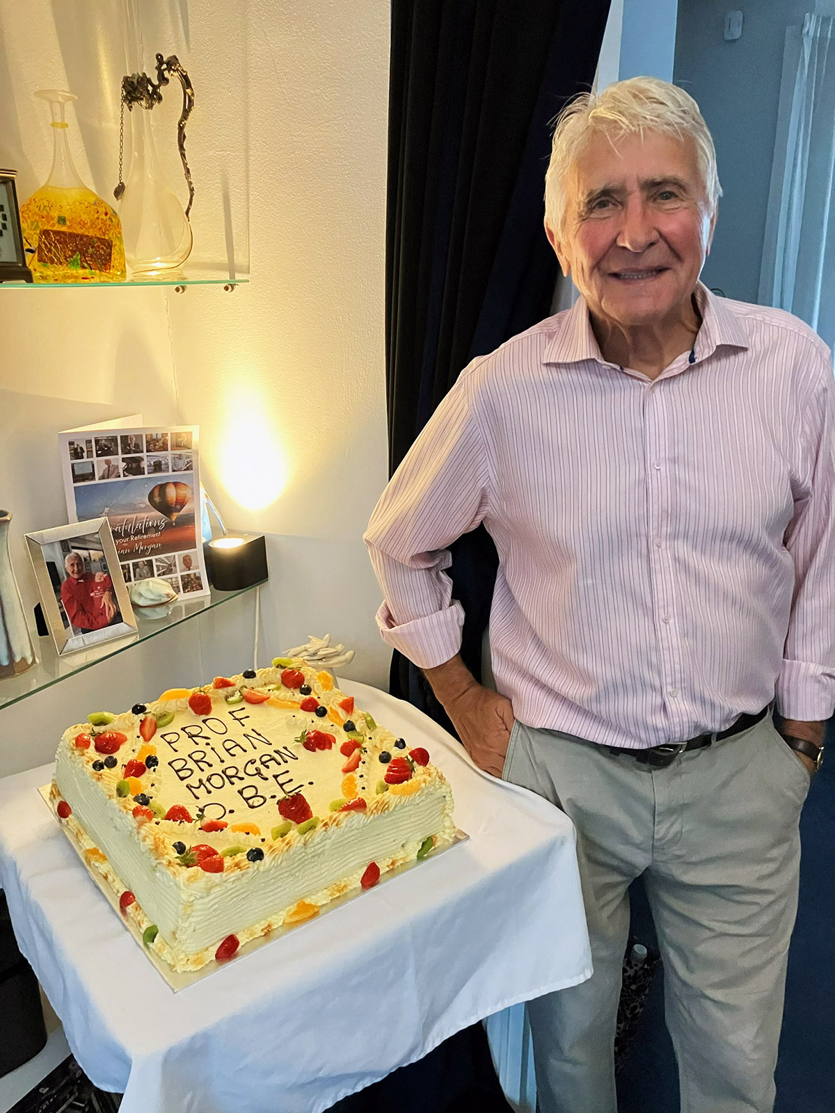 A man stands beside a decorated cake, smiling and celebrating receiving his OBE.