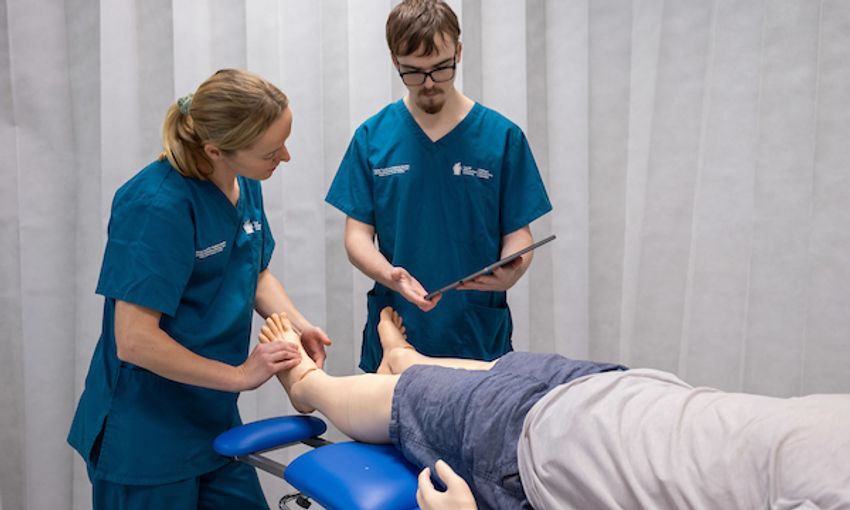 A male and female nurse in blue scrubs consult a chart as they stand at the end of a hospital bed where a test dummy lies