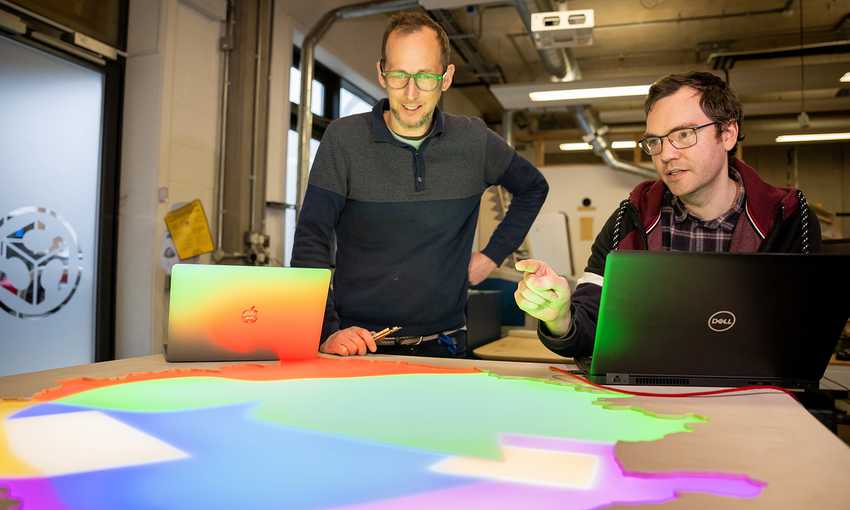 Two men collaborating on a computer, analyzing a colorful light display on the desk in front.