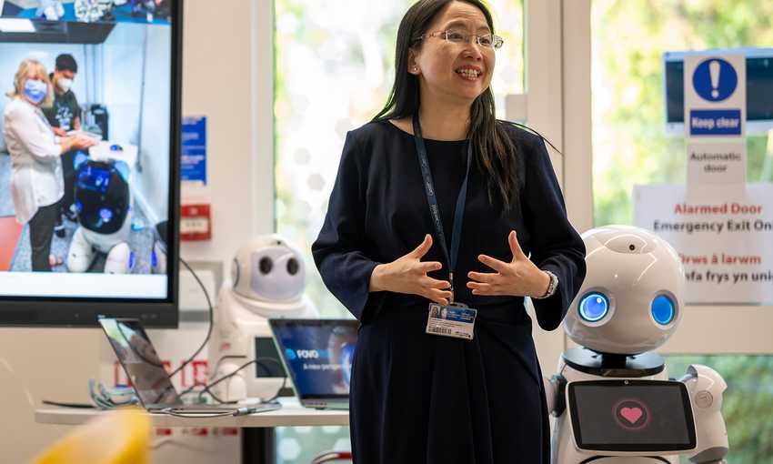 A woman in a black dress stands beside a robot, discussing technology with her students.
