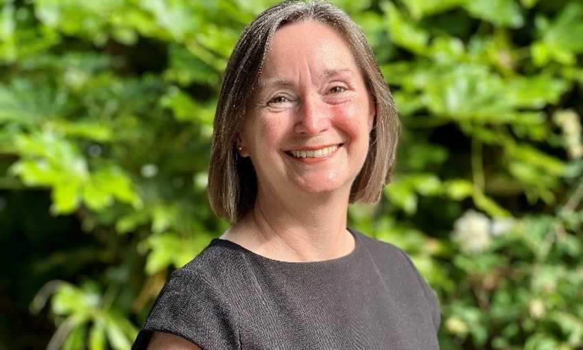A woman in a black top smiles warmly in front of lush green plants, creating a vibrant and cheerful atmosphere.