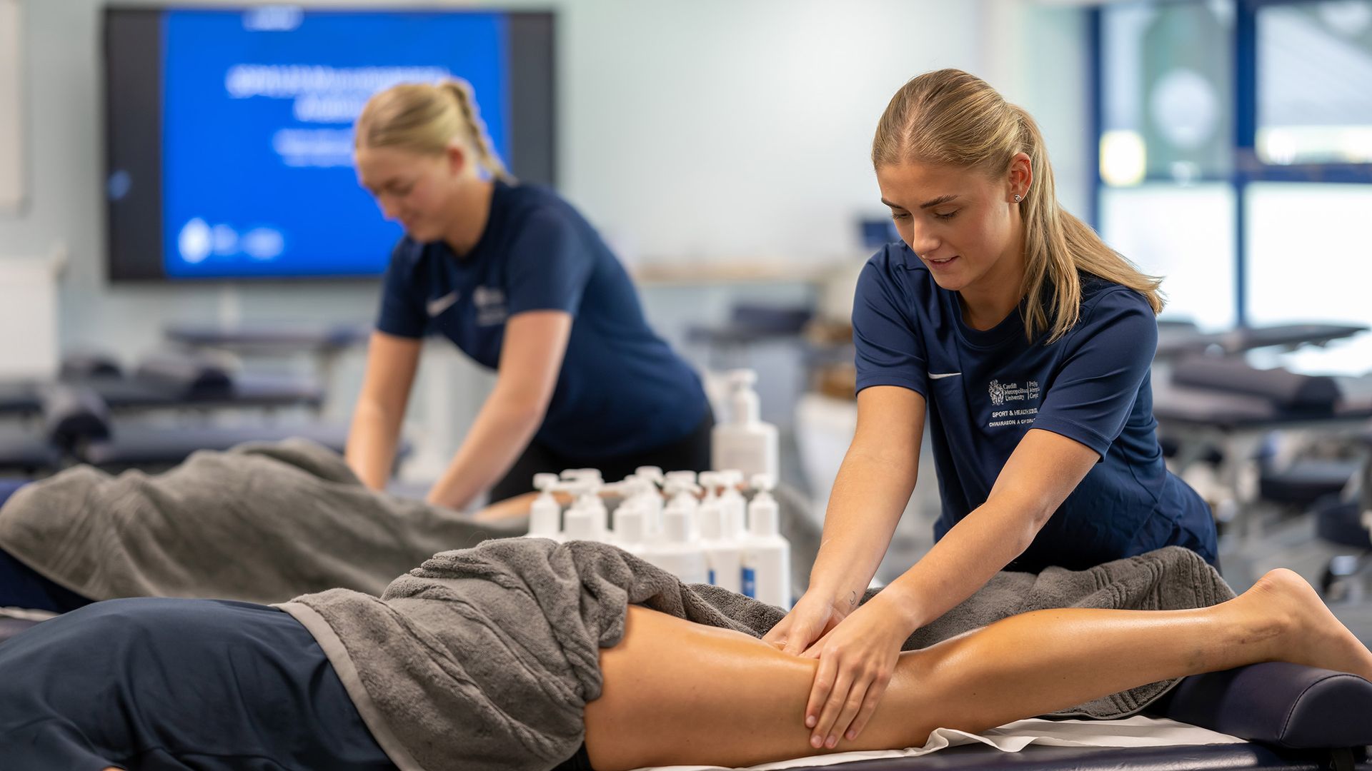 A woman in a navy shirt is smiling as she gives a leg massage to a person lying on a massage table. The setting appears to be a clinic or therapy room with multiple tables and lotion bottles around.
