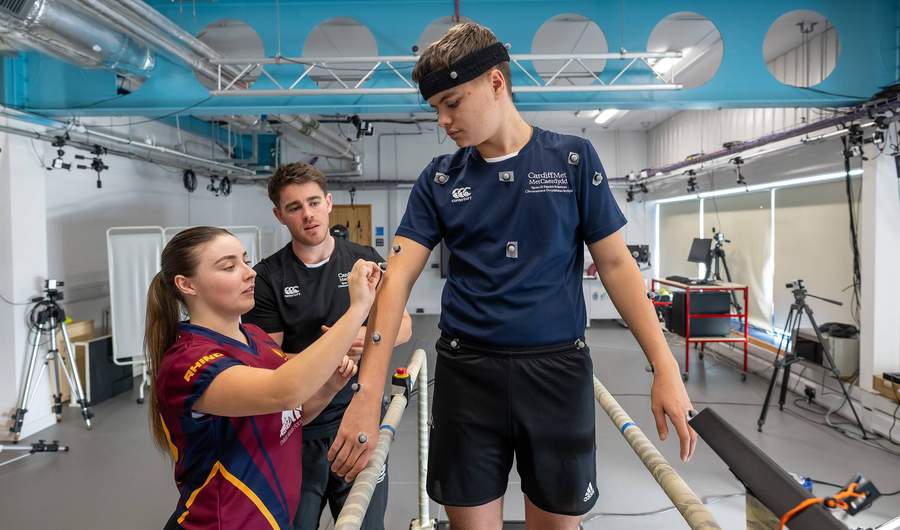 A person stands on a treadmill while another person attaches electrodes to their arms.