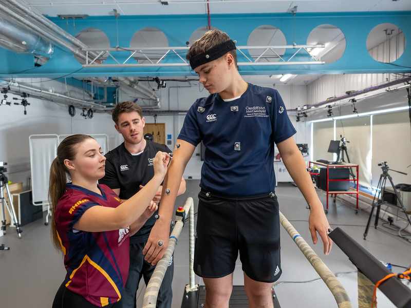 A person stands on a treadmill while another person attaches electrodes to their arms.