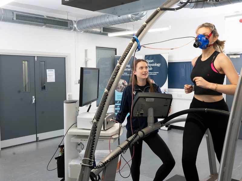 A person runs on a treadmill while wearing a breathing mask. A second person stands alongside, next to a computer display and other equipment.