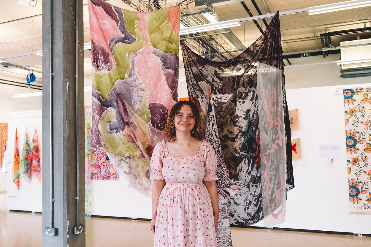 A young woman in a light pink dress stands in front of two large patterned silks