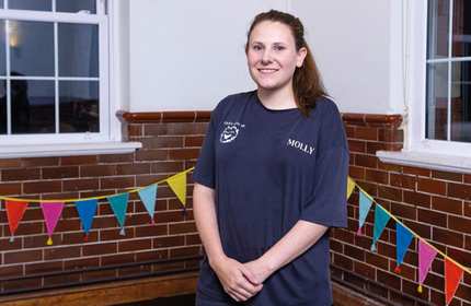 A young woman in a navy blue t-shirt stands in the corner of a tiled and painted room decorated with colourful bunting