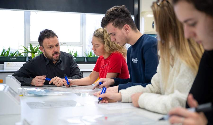 A person kneels next to a table. Four other people sit at the table using marker pens to write.