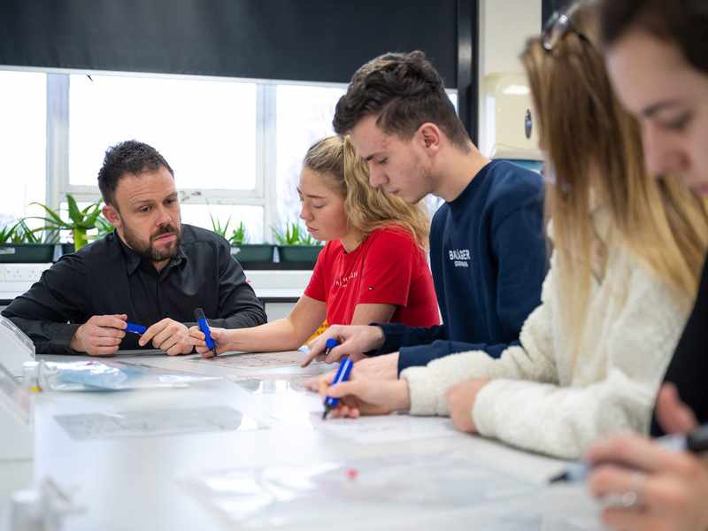 A person kneels next to a table. Four other people sit at the table using marker pens to write.