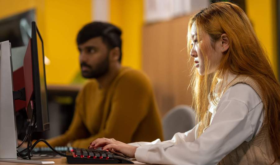 A person working intently at a computer workstation. In the background another person also works at a computer.