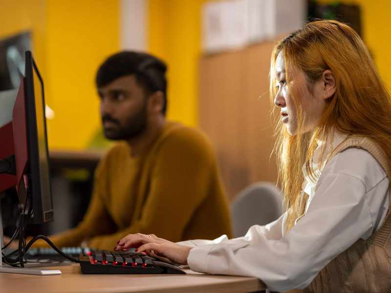A person working intently at a computer workstation. In the background another person also works at a computer.
