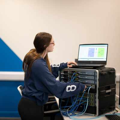 A person plugs cables into computer network equipment while looking at a laptop screen.