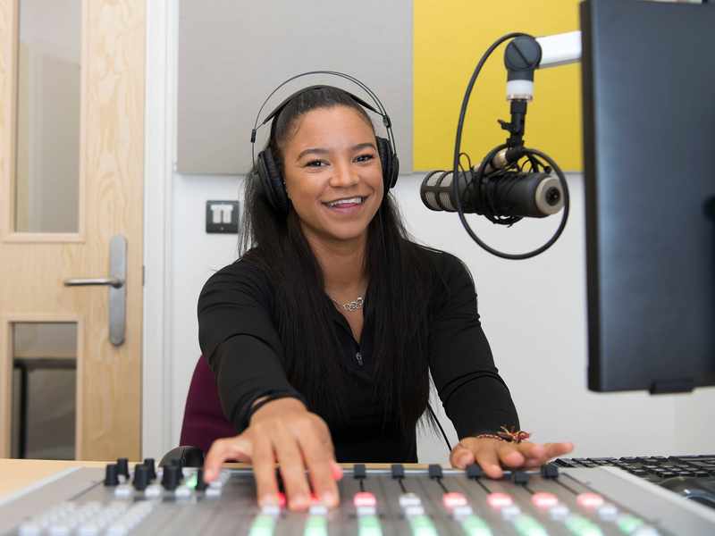 A person wearing headphones sits at a sound mixing desk. In front of them is a microphone mounted on an adjustable arm.