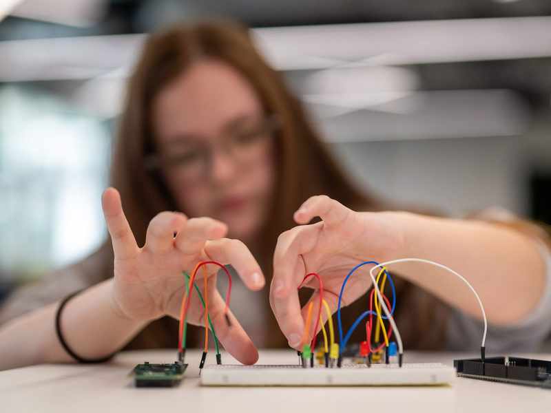 A person with long hair and glasses is working with a breadboard and wires, connecting electronic components. The background is blurred, focusing on the hands and the breadboard.