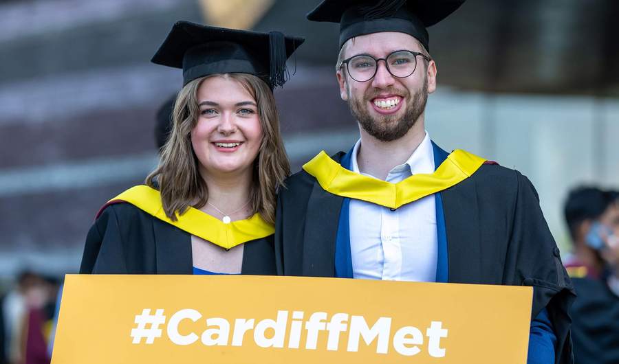 Two people wearing graduation robes hold a sign that reads Cardiff Met.
