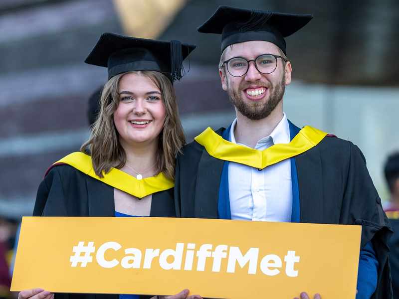 Two people wearing graduation robes hold a sign that reads Cardiff Met.