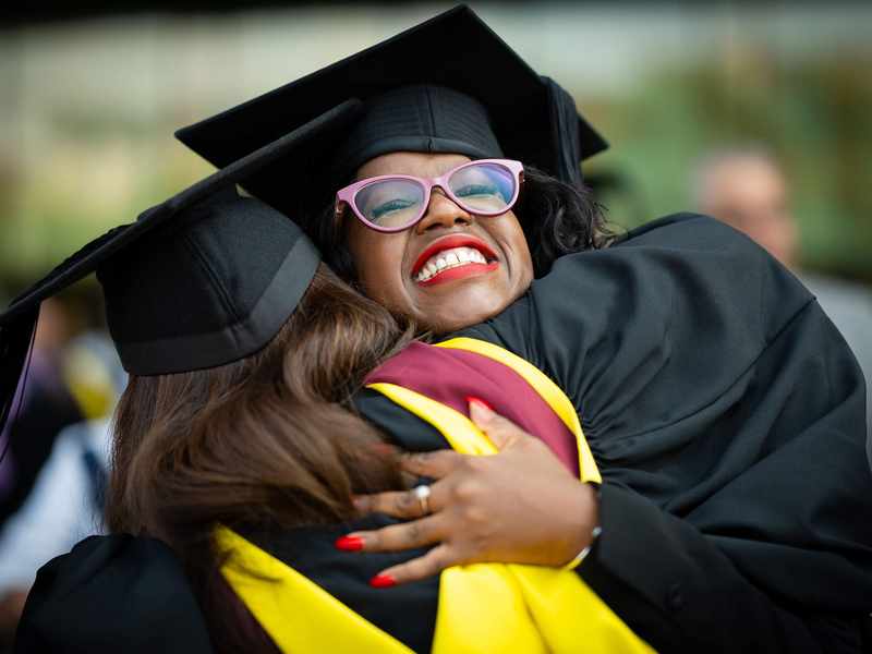 Two students in graduation caps and gowns hug each other.